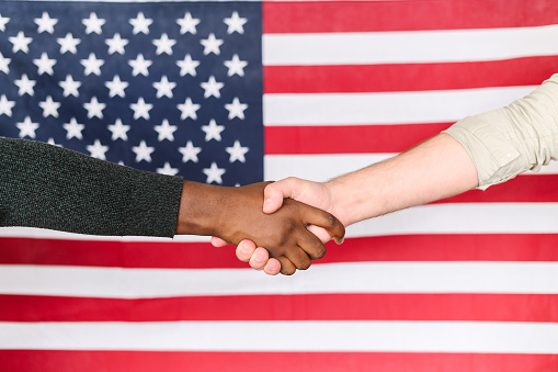 Close-up of black and white males shaking hands with an American flag on the background