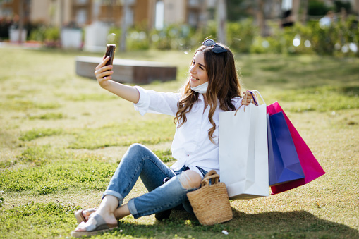 Beautiful young woman shopaholic in medical face mask hold mobile phone and shopping bags in hands. Happy girl in protective face mask shopping online. Coronavirus COVID-19 sale, discount, e-commerce