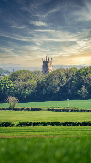 Distant view of the Church of St John the Baptist in Cirencester, Gloucestershire, poking its tower above the surrounding beautiful Cotswold landscape.