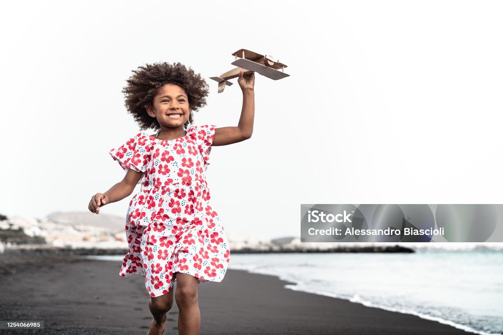 Afro child playing with wood toy airplane on the beach - Little kid having fun during summer holidays - Childhood and travel vacation concept Child Stock Photo