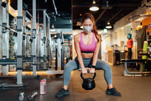 Female athlete wearing protective face mask and lifting kettlebell in gym A female athlete is wearing a protective face mask and lifting a kettlebell in a gym. images of female bodybuilders stock pictures, royalty-free photos & images