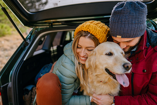 Photo of a young couple in love and their dog sitting the trunk of a car on a beautiful autumn day; taking a short break during their road trip.
