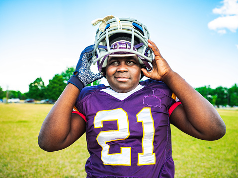 African American Junior Football player getting ready for practice at the outdoor field. He is mounting his protective helmet on.