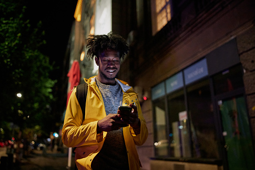Young African American man walking on the street at night, using his mobile phone.