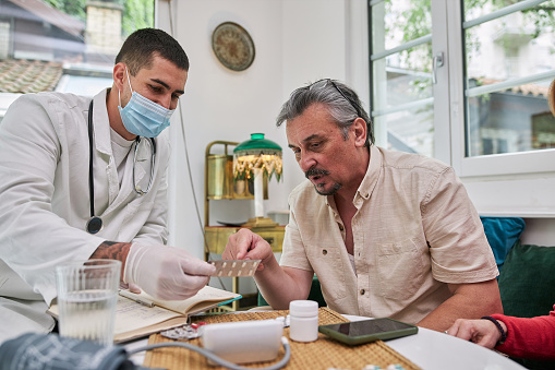 Doctor with a protective face mask visiting a senior man at home, giving him prescription medicine pills, during a coronavirus, covid-19 lockdown.