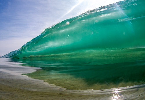 Large clear wave about to break over inches of water with sand getting sucked up from the force of the wave.