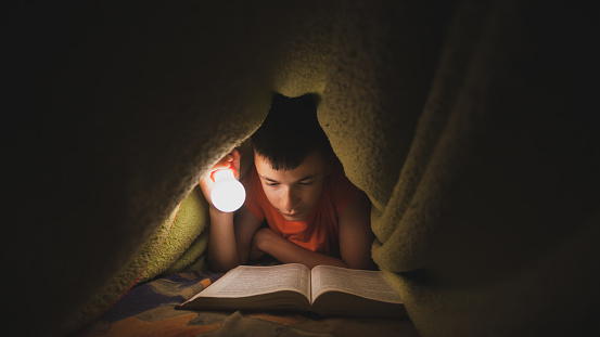 Teenage boy reading a book under the blanket while using a bulb for lightening