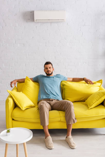 istock Selective focus of handsome man holding remote controller of air conditioner while looking at camera on sofa at home 1254115334
