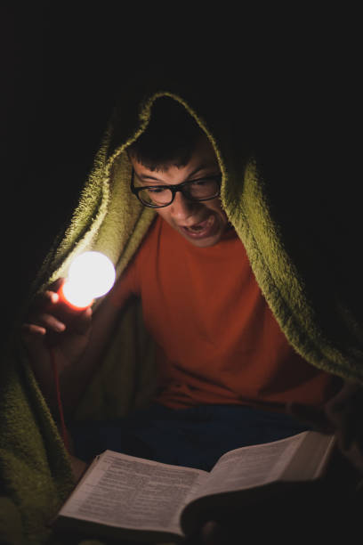 istock Teenage boy reading a book under the blanket at night 1254115564