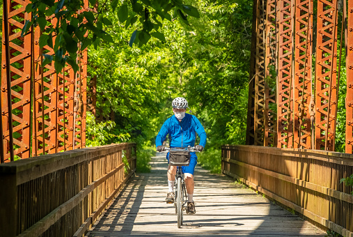 Senior man bicycling along converted railroad bridge on sunny summer day in Midwestern state park wearing a protective face mask; trees in background