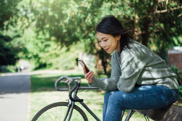 młoda kobieta z inteligentnym telefonem siedzącym na ławce w parku - mental health women asian ethnicity bicycle zdjęcia i obrazy z banku zdjęć