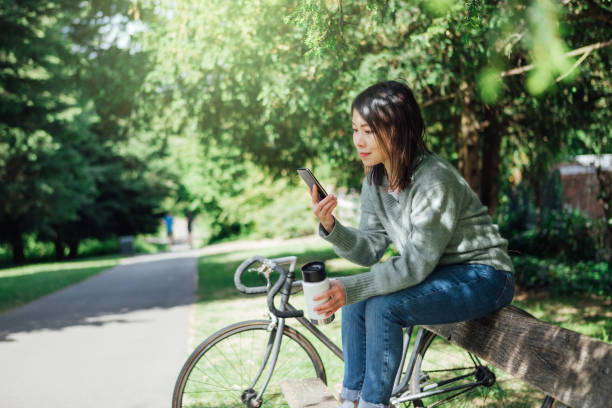 jeune femme avec le téléphone intelligent s’asseyant sur le banc au parc - mental health women asian ethnicity bicycle photos et images de collection