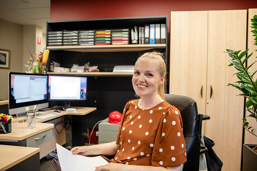 A smiling front view of a woman with muscular dystrophy holding paperwork smiling and looking into the camera at work in the office in Perth, Western Australia.