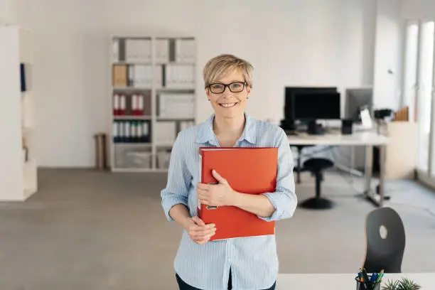 Smiling happy businesswoman clutching a large red binder to her chest as she stands in a spacious modern office