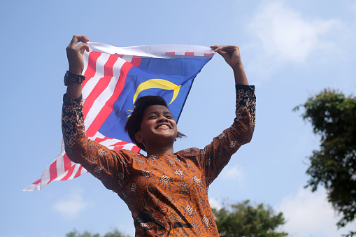 Muslim Indian ethnicity teenage girl is enjoying nature reserve environment while holding Malaysia flag cheerfully.