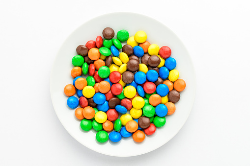 Group of small red, yellow, green, blue, brown and orange coated chocolate candies in a white round plate isolated on white background, top view