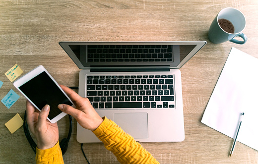 Close-up on a woman using app on her cell phone while working online at home on her laptop computer