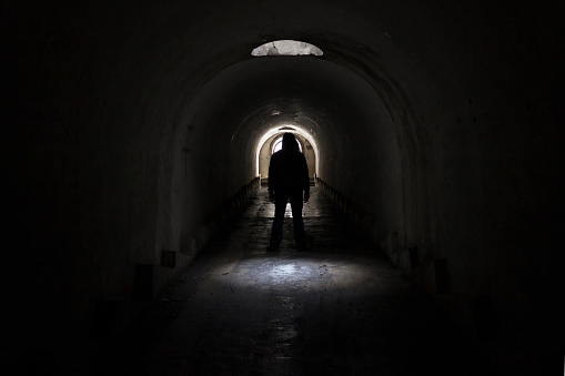A silhouette of a man in dark clothes with a hood going to the exit to the lighted door from a dark terrible underground passage lit through a hatch in the ceiling.