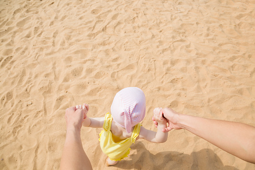 Young mother helping baby girl taking first steps at beach. Lovely, peaceful walking together on sand in sunny summer day. Point of view shot. Empty place for text. Top down view.