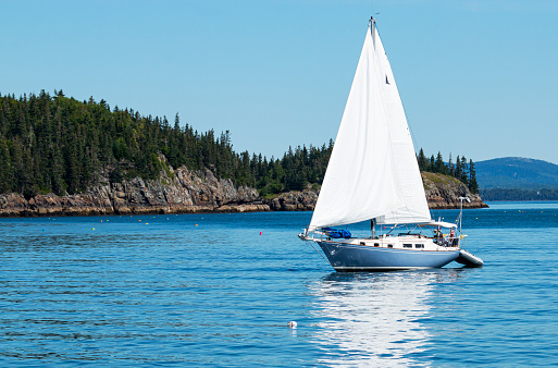 A light blue sailboat is sailing peacefully in Frenchman Bay around the porcupine islands in Bar Harbor Maine.