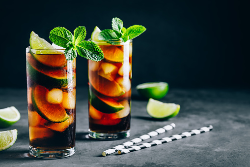 Rum and Cola Cuba Libre ice cold drink cocktail with lime and mint on dark background.