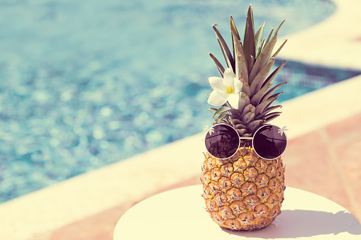 Funny happy pineapple in sunglasses on swimming pool background at tropical sunny day. Creative food and travel concept card with hipster ananas in glasses