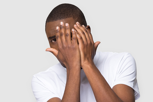 Scared unhappy African American man hiding behind hands, frightened young male looking at camera, feeling afraid, stress, negative emotion, horror movie, problem, isolated on studio background