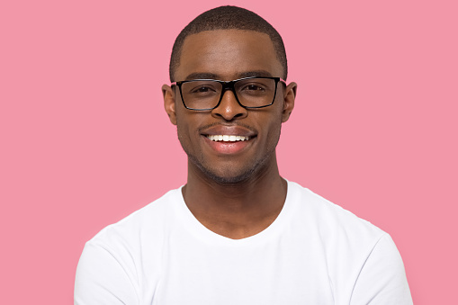 Head shot portrait African American man in glasses looking at camera, satisfied male with healthy toothy smile, satisfied teenage student in white t-shirt, isolated on pink studio background