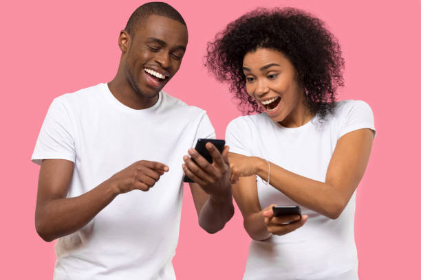 Excited African American couple using mobile phone, reading good news Excited African American couple using mobile phone, smiling laughing man and woman reading good news in social network or email, great shopping offer, online win, isolated on pink studio background unusual couple stock pictures, royalty-free photos & images