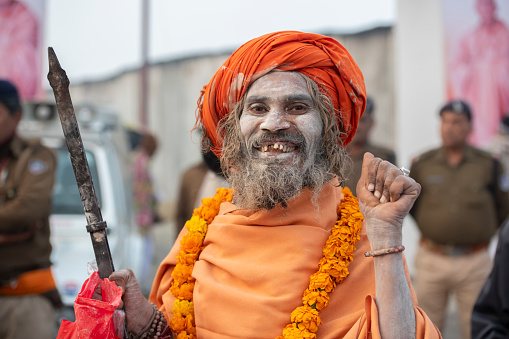 Portrait of a very friendly and smiling sadhu (holy man), wearing orange turban, mantle and garland, while holding his tongs. They are a symbol of the fire they keep burning for their worship of Shiva. This man vibrated as an electric coil. Photo taken during the Royal bath procession in Kumbh Mela 2019, Prayagraj (Allahabad), India. \nKumbh Mela or Kumbha Mela is a major pilgrimage and festival in Hinduism, and probably the greatest religious festival in the World. It is celebrated in a cycle of approximately 12 years at four river-bank pilgrimage sites: the Allahabad (Ganges-Yamuna Sarasvati rivers confluence), Haridwar (Ganges), Nashik (Godavari), and Ujjain (Shipra). The festival is marked by a ritual dip in the waters, but it is also a celebration of community commerce with numerous fairs, education, religious discourses by saints, mass feedings of monks or the poor, and entertainment spectacles. Pilgrims believe that bathing in these rivers is a means to cleanse them of their sins and favour a better next incarnation.