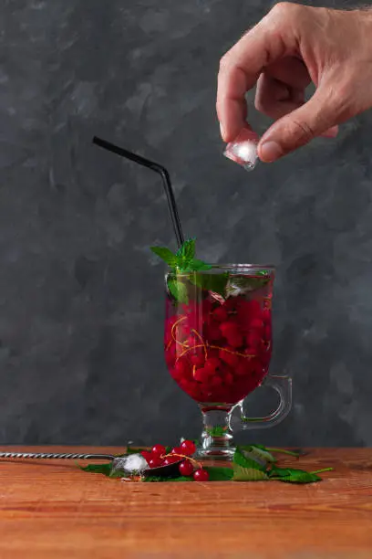 Summer red currant tea in a glass on dark background. Male hand holds melting ice cube with drop over fruit cold cocktail. Tasty iced juice with fresh green leaves.Seasonal vitamins healthy food photo