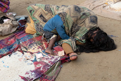 Leper woman, with the fingers missing from the left hand, laying on the floor. A child is holding to her, hiding under a piece of clothing. A bag and shoes lay near her feet, while rice, small coins and bills are shown in her front. \nDespite leprosy being comparatively cheap to cure, these persons are kept sick by gangs who explore them. They are left in strategic places, by the hundreds, so people can give alms in hope for a good karma, after taking the holy dip.\nPhoto taken during Kumbh Mela 2019 in Prayagraj (Allahabad), India. \nKumbh Mela or Kumbha Mela is a major pilgrimage and festival in Hinduism, and probably the greatest religious festival in the World. It is celebrated in a cycle of approximately 12 years at four river-bank pilgrimage sites: the Allahabad (Ganges-Yamuna Sarasvati rivers confluence), Haridwar (Ganges), Nashik (Godavari), and Ujjain (Shipra). The festival is marked by a ritual dip in the waters, but it is also a celebration of community commerce with numerous fairs, education, religious discourses by saints, mass feedings of monks or the poor, and entertainment spectacles. Pilgrims believe that bathing in these rivers is a means to cleanse them of their sins and favour a better next incarnation.