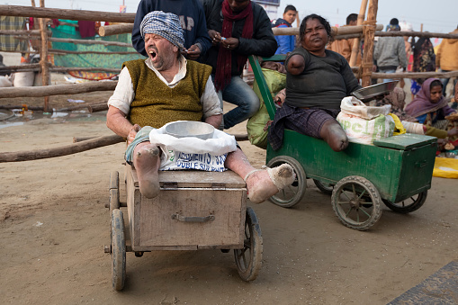 Two amputee lepers, man and woman on wooden carts, being displayed for profit among a crowd of beggars. They also seem to suffer from some sort of mental illness.  Despite leprosy being comparatively cheap to cure, these persons are kept sick by gangs who explore them.  They are left in strategic places, by the hundreds, so people can give alms in hope for a good karma, after taking the holy dip.Photo taken during Kumbh Mela 2019 in Prayagraj (Allahabad), India. \nKumbh Mela or Kumbha Mela is a major pilgrimage and festival in Hinduism, and probably the greatest religious festival in the World. It is celebrated in a cycle of approximately 12 years at four river-bank pilgrimage sites: the Allahabad (Ganges-Yamuna Sarasvati rivers confluence), Haridwar (Ganges), Nashik (Godavari), and Ujjain (Shipra). The festival is marked by a ritual dip in the waters, but it is also a celebration of community commerce with numerous fairs, education, religious discourses by saints, mass feedings of monks or the poor, and entertainment spectacles. Pilgrims believe that bathing in these rivers is a means to cleanses them of their sins and favour a better next incarnation.