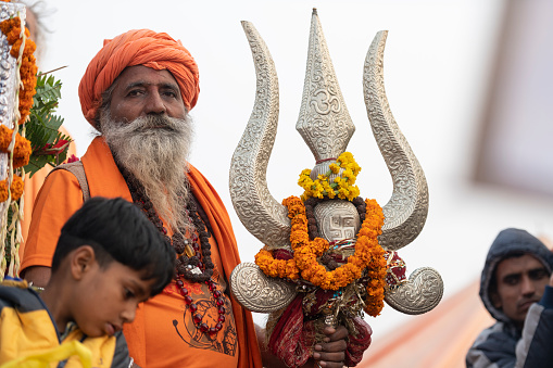Holy man, wearing orange turban and t-shirt, holding a large, silver Trishula, wrapped in garlands. He's on a float, along with a young boy, parading at the Royal Bath procession. Photo taken during Kumbh Mela 2019 in Prayagraj (Allahabad), India. \nTrishulas (tridents) have a wide symbolism: is wielded by the god Shiva and is said to have been used to sever the original head of Ganesha. Durga also holds a trishula, as one of her many weapons. The three points represent various trinities, regarding deities, qualities and tenses. \nKumbh Mela or Kumbha Mela is a major pilgrimage and festival in Hinduism, and probably the greatest religious festival in the World. It is celebrated in a cycle of approximately 12 years at four river-bank pilgrimage sites: the Allahabad (Ganges-Yamuna Sarasvati rivers confluence), Haridwar (Ganges), Nashik (Godavari), and Ujjain (Shipra). The festival is marked by a ritual dip in the waters, but it is also a celebration of community commerce with numerous fairs, education, religious discourses by saints, mass feedings of monks or the poor, and entertainment spectacles. Pilgrims believe that bathing in these rivers is a means to cleanse them of their sins and favour a better next incarnation.