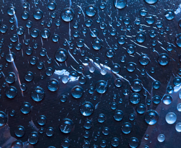 istock Transparent raindrops flow down on the glass  surface of a deep blue background and are lit by the rays of the bright sun. Close-up. 1254233193