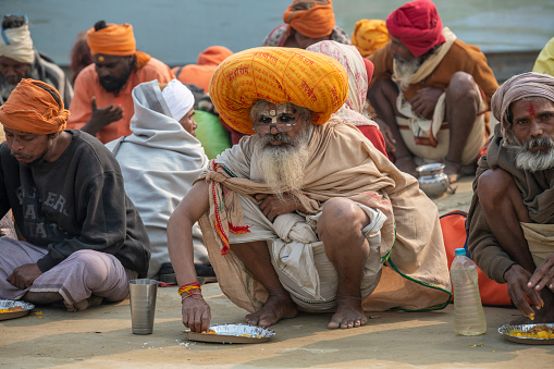 Men, sitting on the floor and eating what seems to me lentil curry during a community meal. It is taking place at one of the busiest Tent-City. Mass feedings of the saints and poor are common in Kumbh Mela festivities, either sponsored by temples or by rich men. Photo taken in 2019, Prayagraj (Allahabad), India. \nKumbh Mela or Kumbha Mela is a major pilgrimage and festival in Hinduism, and probably the greatest religious festival in the World. It is celebrated in a cycle of approximately 12 years at four river-bank pilgrimage sites: the Allahabad (Ganges-Yamuna Sarasvati rivers confluence), Haridwar (Ganges), Nashik (Godavari), and Ujjain (Shipra). The festival is marked by a ritual dip in the waters, but it is also a celebration of community commerce with numerous fairs, education, religious discourses by saints, and entertainment spectacles. Pilgrims believe that bathing in these rivers is a means to cleanse them of their sins and favour a better next incarnation.