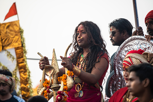 Hijra holding a brass Trisula (trident) while on a float, parading in the Royal Bath procession. Photo taken during Kumbh Mela 2019 in Prayagraj (Allahabad), India. \nTrishulas (tridents) have a wide symbolism: is wielded by the god Shiva and is said to have been used to sever the original head of Ganesha. Durga also holds a trishula, as one of her many weapons. The three points represent various trinities, regarding deities, qualities and tenses.\nHijra are eunuchs, intersex and transgender people. They are gathered in an organization (Kinnar Akhada) and for the first time, they were allowed to set their own camp in the tent city, and had a major role on the processions. \nKumbh Mela or Kumbha Mela is a major pilgrimage and festival in Hinduism, and probably the greatest religious festival in the World. It is celebrated in a cycle of approximately 12 years at four river-bank pilgrimage sites: the Allahabad (Ganges-Yamuna Sarasvati rivers confluence), Haridwar (Ganges), Nashik (Godavari), and Ujjain (Shipra). The festival is marked by a ritual dip in the waters, but it is also a celebration of community commerce with numerous fairs, education, religious discourses by saints, mass feedings of monks or the poor, and entertainment spectacles. Pilgrims believe that bathing in these rivers is a means to cleanse them of their sins and favour a better next incarnation.