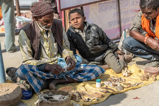 Elderly Indian man sitting on the  floor before a collection of what seems stones and bones. He is accompanied by a teenager, while another man observes the products on display. The collection is either ingredients for traditional medicine, or lucky charms. Photo taken during Kumbh Mela 2019 in Prayagraj (Allahabad), India. \nKumbh Mela or Kumbha Mela is a major pilgrimage and festival in Hinduism, and probably the greatest religious festival in the World. It is celebrated in a cycle of approximately 12 years at four river-bank pilgrimage sites: the Allahabad (Ganges-Yamuna Sarasvati rivers confluence), Haridwar (Ganges), Nashik (Godavari), and Ujjain (Shipra). The festival is marked by a ritual dip in the waters, but it is also a celebration of community commerce with numerous fairs, education, religious discourses by saints, mass feedings of monks or the poor, and entertainment spectacles. Pilgrims believe that bathing in these rivers is a means to cleanse them of their sins and favour a better next incarnation.