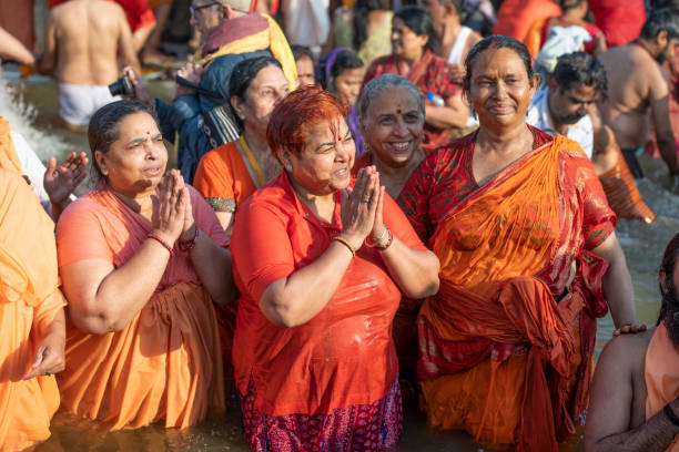 Indian women devotees, wearing red and orange, clasping hands and praying. They are taking the holy dip, surrounded by a crowd, in the Royal bath day. Photo taken during Kumbh Mela 2019 in Prayagraj (Allahabad), India. \nKumbh Mela or Kumbha Mela is a major pilgrimage and festival in Hinduism, and probably the greatest religious festival in the World. It is celebrated in a cycle of approximately 12 years at four river-bank pilgrimage sites: the Allahabad (Ganges-Yamuna Sarasvati rivers confluence), Haridwar (Ganges), Nashik (Godavari), and Ujjain (Shipra). The festival is marked by a ritual dip in the waters, but it is also a celebration of community commerce with numerous fairs, education, religious discourses by saints, mass feedings of monks or the poor, and entertainment spectacles. Pilgrims believe that bathing in these rivers is a means to cleanse them of their sins and favour a better next incarnation.