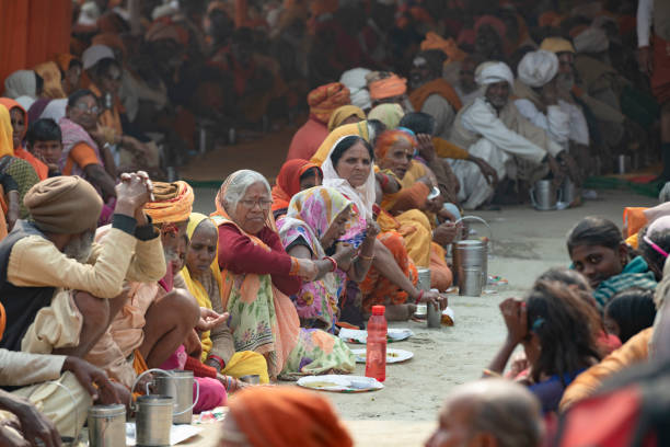 Poor people sitting on the floor during a mass feeding, or community meal, in a tent. Mass feedings of the saints and poor are common in Kumbh Mela festivities, either sponsored by temples or by rich men. Photo taken in 2019, Prayagraj (Allahabad), India. \nKumbh Mela or Kumbha Mela is a major pilgrimage and festival in Hinduism, and probably the greatest religious festival in the World. It is celebrated in a cycle of approximately 12 years at four river-bank pilgrimage sites: the Allahabad (Ganges-Yamuna Sarasvati rivers confluence), Haridwar (Ganges), Nashik (Godavari), and Ujjain (Shipra). The festival is marked by a ritual dip in the waters, but it is also a celebration of community commerce with numerous fairs, education, religious discourses by saints, and entertainment spectacles. Pilgrims believe that bathing in these rivers is a means to cleanse them of their sins and favour a better next incarnation.