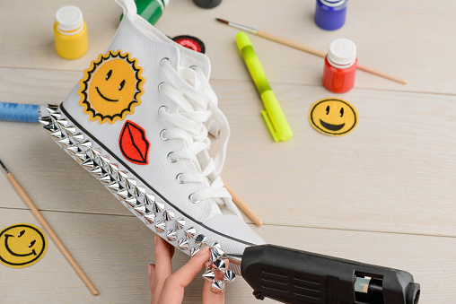 Girl putting metal spikes onto the sole of sneakers using a hot glue gun. Teenage fashion and handmade projects.
