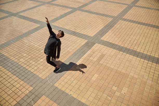 Handsome guy in black outfit raising hand and looking up while standing on concrete sidewalk on sunny day