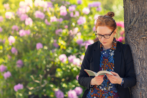 A woman standing by a tree and reading a book in a garden with blooming rhododendron bushes.