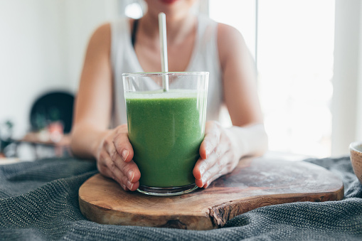 Cropped and close-up shot of young woman holding and drinking a glass of green smoothie for breakfast.