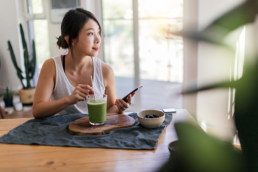 Medium shot of young Asian woman using smart phone while drinking green smoothie and eating fruits and nuts for breakfast in the morning at home.