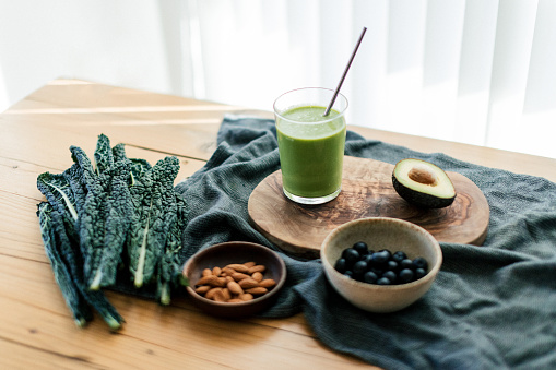 A glass of green smoothie with metal straw on the table with raw vegan food and ingredients.