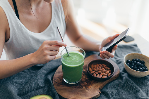 Close-up shot of young woman using smart phone while drinking green smoothie and eating fruits and nuts for breakfast in the morning at home.