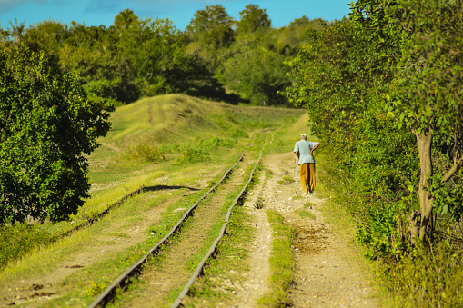 An elderly Cuban man walks with a walking stick next to a train line in the Cuban countryside