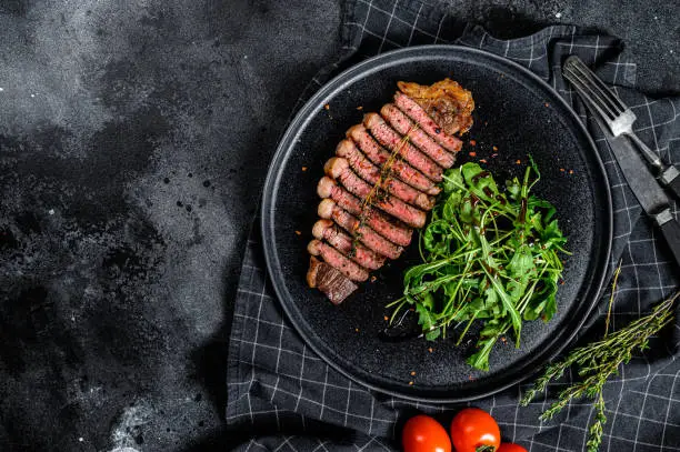 Sliced sirloin steak, marbled beef meat with arugula. Black background. Top view. Copy space.