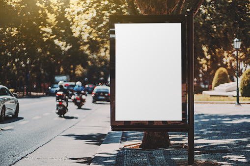 Blank white advert billboard template on a paving stone near the road; empty ad banner placeholder mockup template on the sidewalk; poster mock-up in urban settings in the shadow of an alleyway